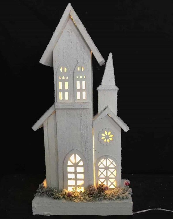 Tall white wooden Christmas chapel with illuminated windows, steeple, snowy finish, greenery and pinecone accents around the base.